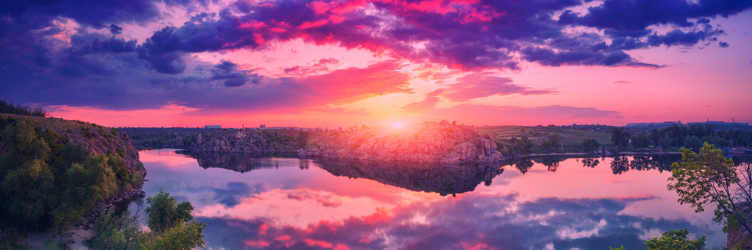 Vibrant sunset over a calm lake surrounded by rocky cliffs and trees, with dramatic purple and pink clouds reflecting on the water.